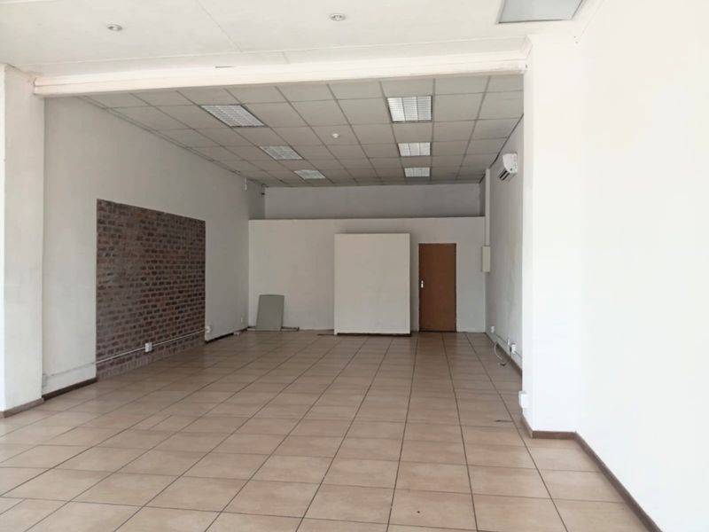 A spacious, empty open-plan retail unit featuring tiled flooring, a suspended ceiling with recessed lighting, and an exposed brick feature wall.