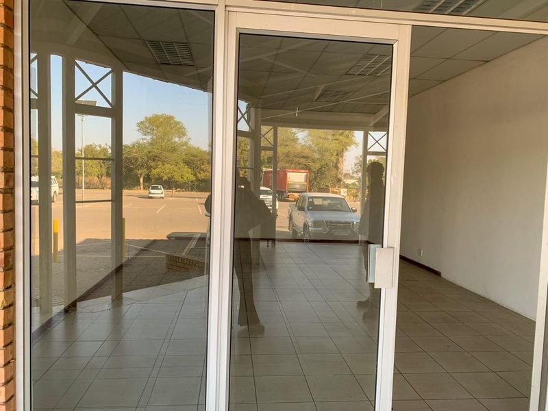 A bright, vacant commercial entrance area featuring tiled flooring, a suspended ceiling, and large glass doors providing clear visibility to the exterior parking area.