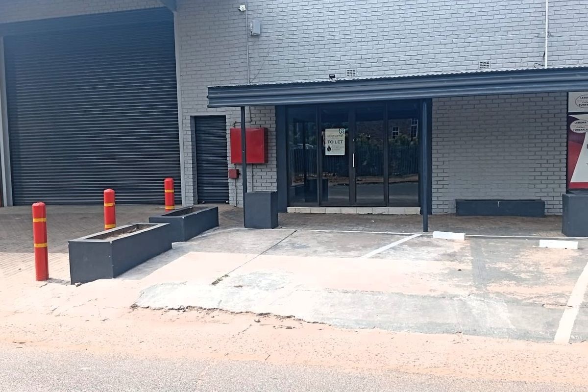 Front exterior view of an office or industrial unit featuring a roller shutter door, glass entrance, and dedicated concrete parking area with bollards.