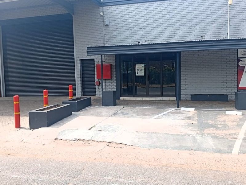 Front exterior view of an office or industrial unit featuring a roller shutter door, glass entrance, and dedicated concrete parking area with bollards.