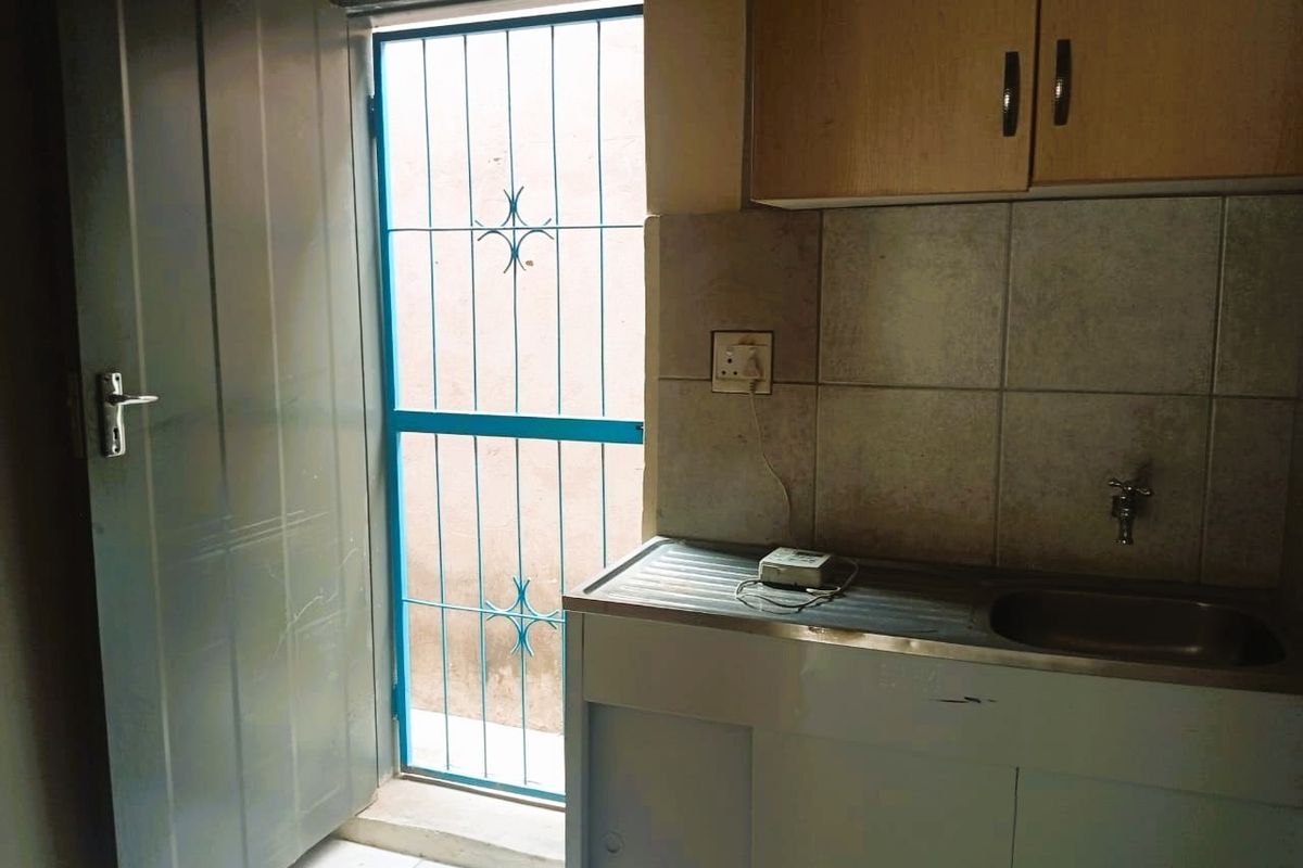 A compact kitchen area featuring a stainless steel sink, tiled backsplash, and built-in cabinetry, with direct access to an exterior security gate.
