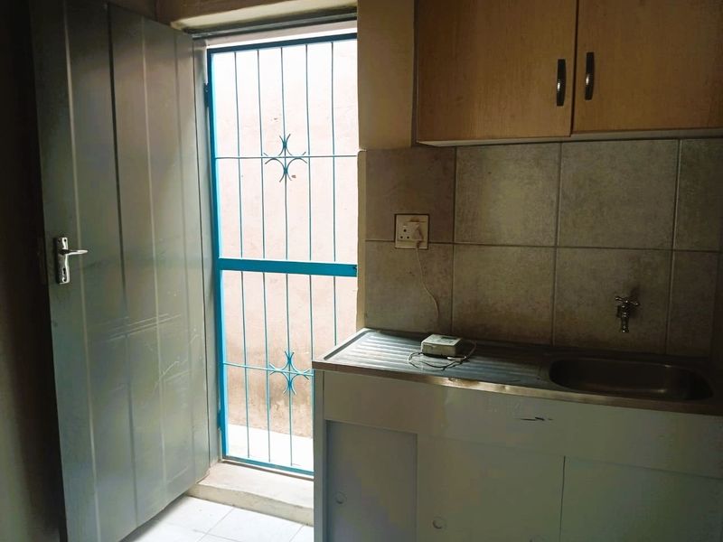 A compact kitchen area featuring a stainless steel sink, tiled backsplash, and built-in cabinetry, with direct access to an exterior security gate.