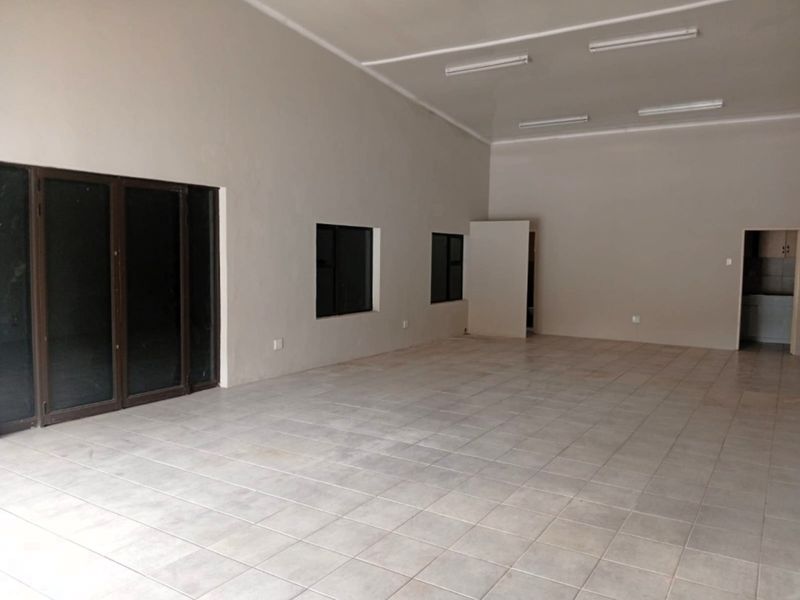 A spacious, empty office area featuring neutral tiled flooring, large sliding glass doors, and functional overhead fluorescent lighting.