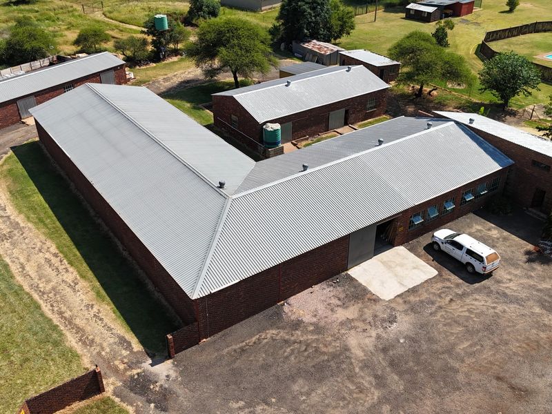 Aerial view of a small holding property featuring multiple brick outbuildings with corrugated metal roofs, a swimming pool, and expansive grounds.