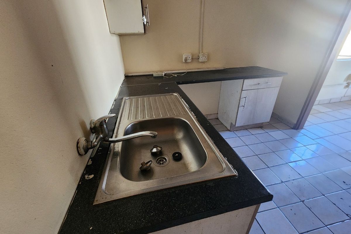 A basic kitchenette area featuring a stainless steel sink, laminate countertops, and tiled flooring, currently in need of cosmetic updates and maintenance.