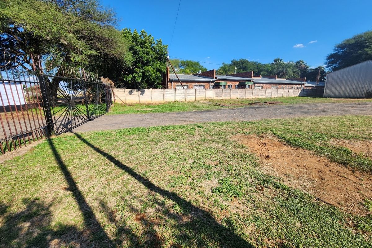 A view of the industrial property entrance featuring a metal security gate with razor wire, a paved driveway, and perimeter precast concrete walling.
