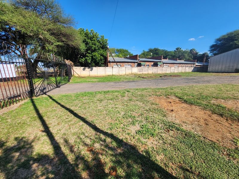 A view of the industrial property entrance featuring a metal security gate with razor wire, a paved driveway, and perimeter precast concrete walling.