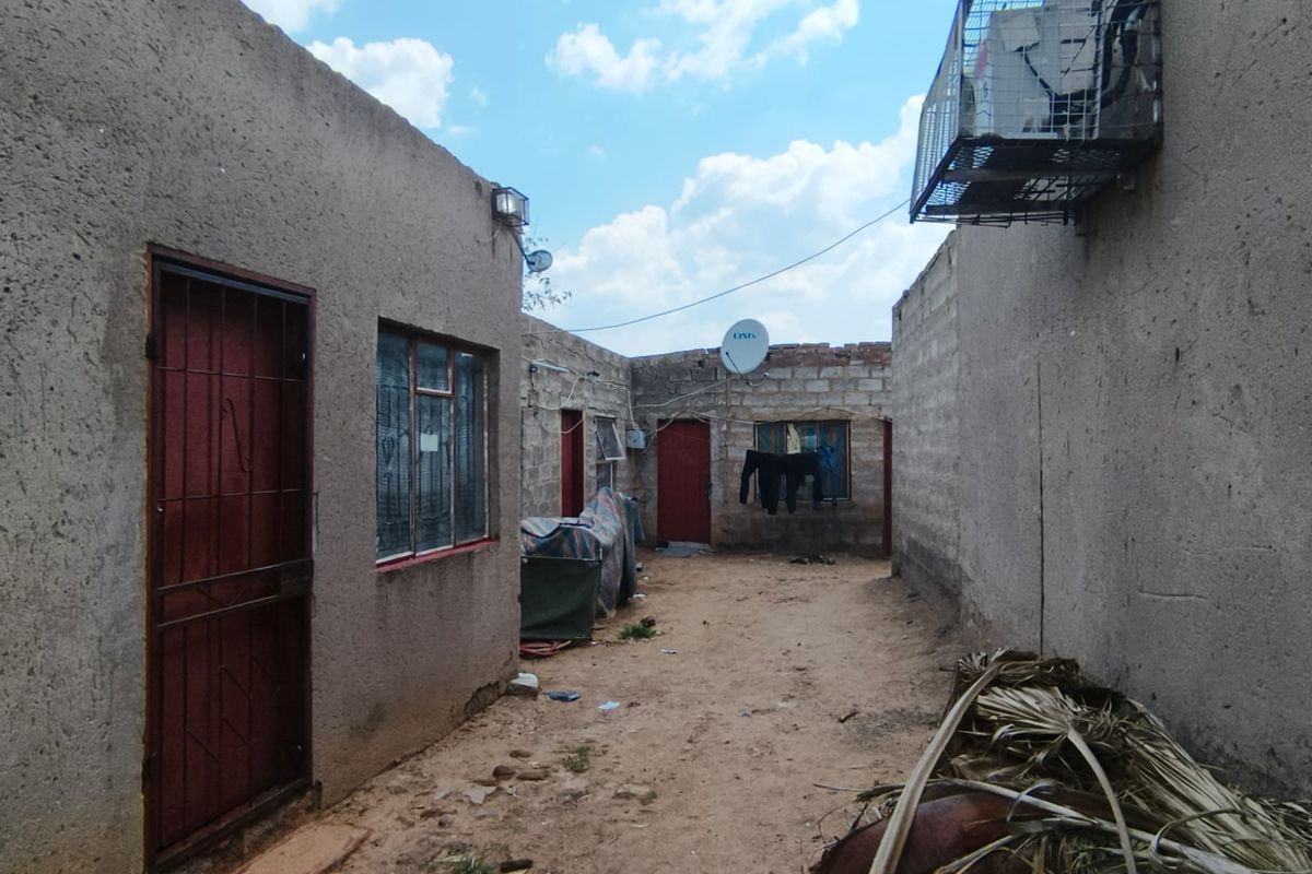 Rear exterior view of the property showing multiple access points, security gates, and unpaved yard space with exposed brick and plaster finishes.
