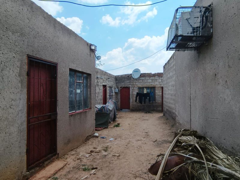 Rear exterior view of the property showing multiple access points, security gates, and unpaved yard space with exposed brick and plaster finishes.