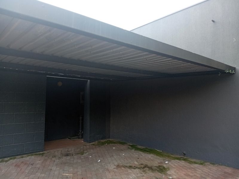 A covered exterior entrance area featuring a metal carport structure, brick paved flooring, and dark-painted concrete block walls.