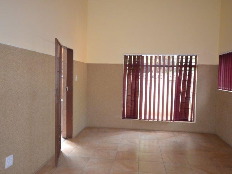 An empty, neutrally painted living area featuring tiled flooring, textured wall finishes, and security-fitted windows with vertical blinds.