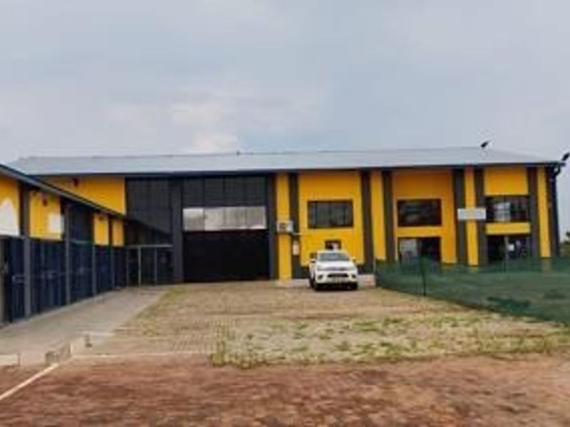 Front elevation of an industrial warehouse facility featuring a large roller shutter door, paved driveway, and metal roof structure.