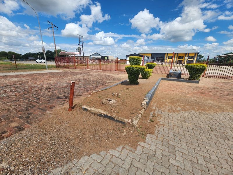 Exterior view of an industrial property featuring a paved driveway, metal perimeter fencing, and a raised garden bed area requiring maintenance.