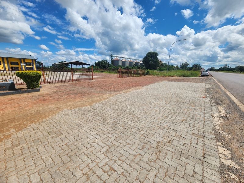 Spacious industrial exterior area featuring extensive paved driveway, metal perimeter fencing, and a covered parking structure with distant industrial storage silos.