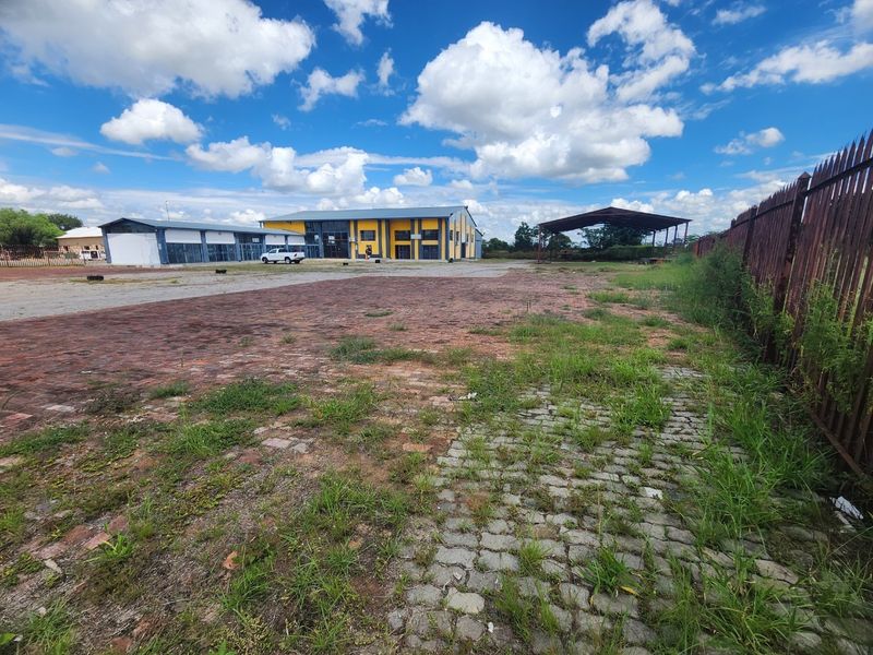 Expansive industrial yard featuring brick and cobblestone paving, perimeter fencing, and access to a large warehouse building and an open-sided metal storage shed.