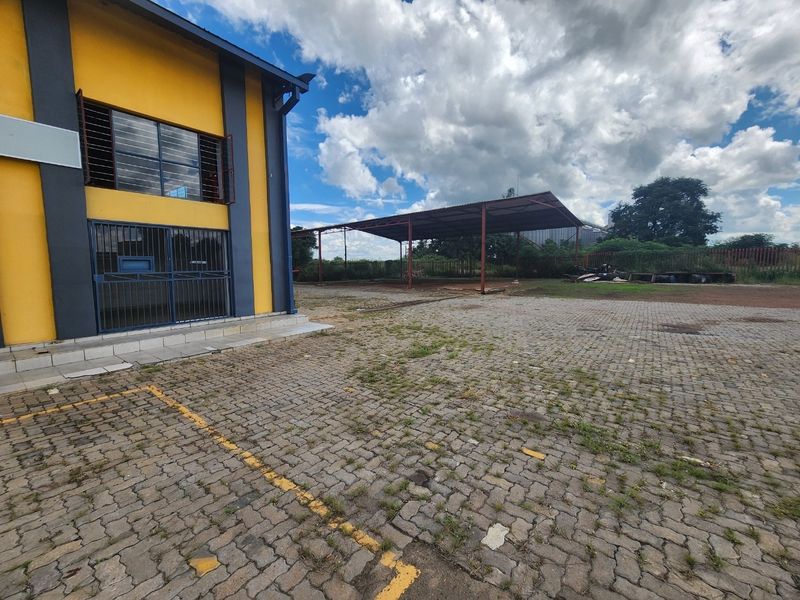 Industrial property exterior featuring a paved yard area, security-fitted windows, and a large steel-framed covered structure for storage or vehicle access.