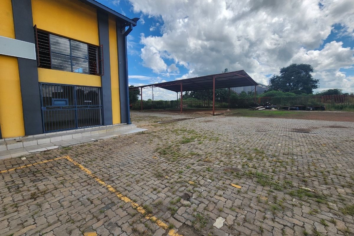Industrial property exterior featuring a paved yard area, security-fitted windows, and a large steel-framed covered structure for storage or vehicle access.