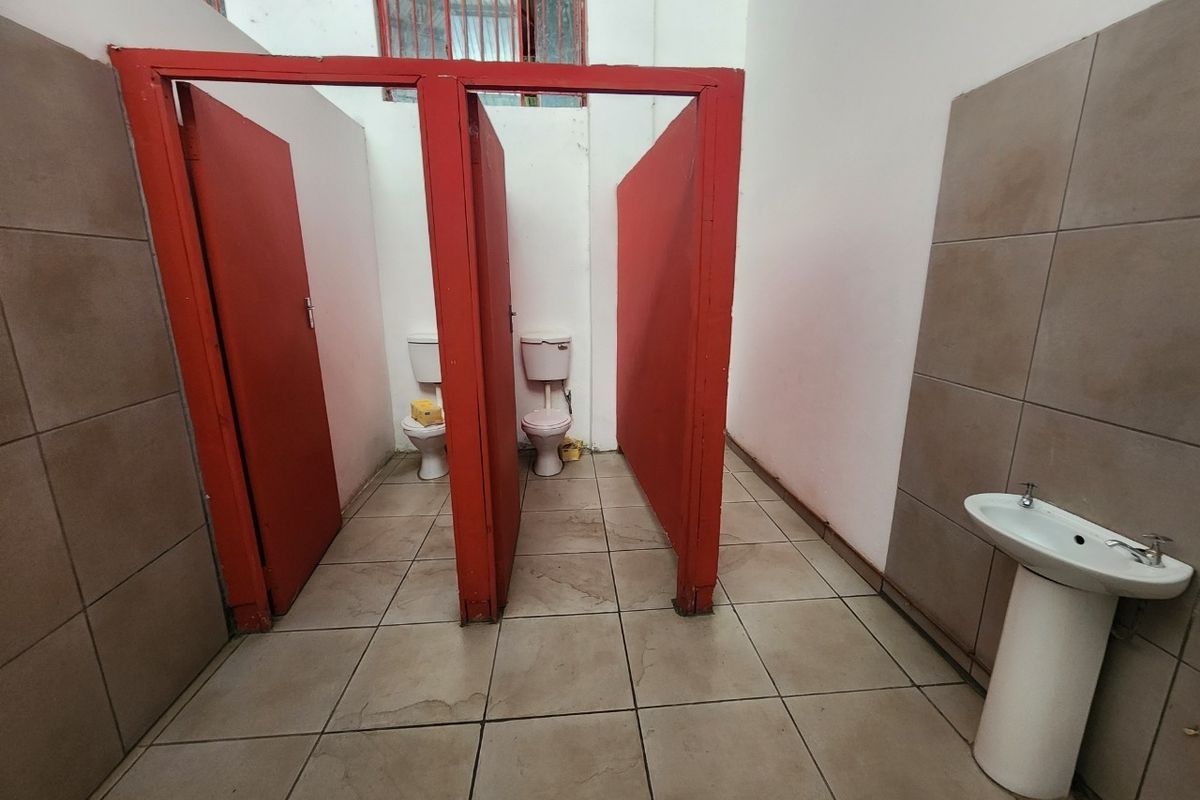 Functional industrial-style communal bathroom featuring two toilet cubicles with red partitions and a single pedestal basin with tiled flooring and wall accents.