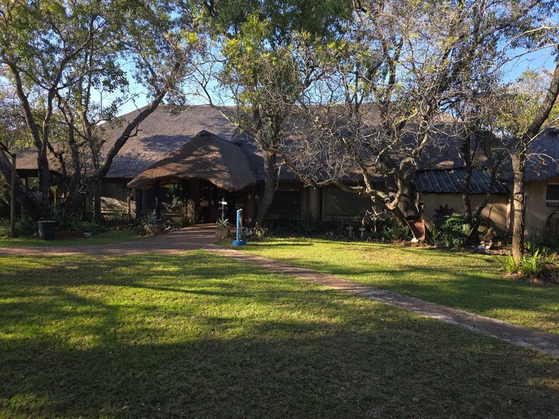 Front view of a farm-style residence featuring a traditional thatched roof, surrounded by mature trees and a well-maintained lawn with a paved entrance path.