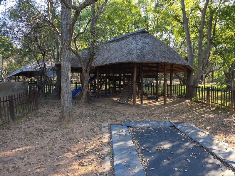 A rustic outdoor garden area featuring a large thatched gazebo structure, timber fencing, and an in-ground trampoline set within a wooded landscape.