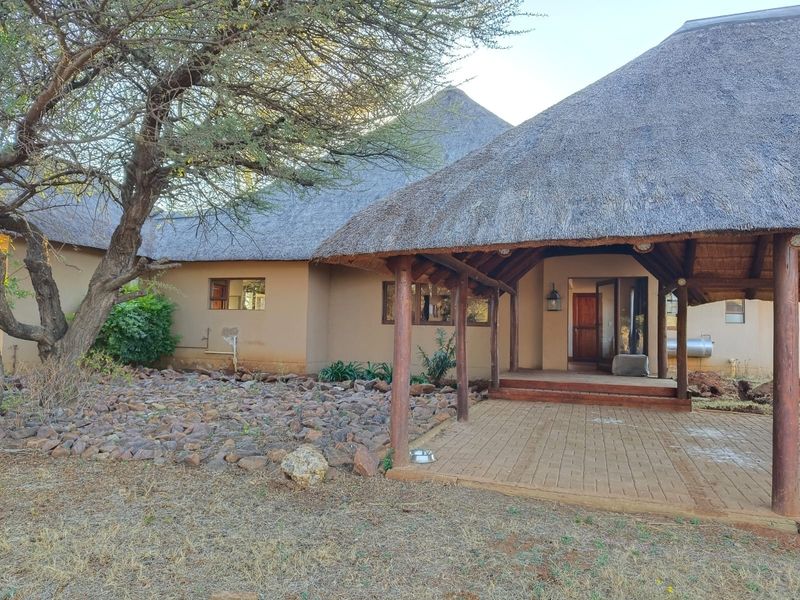 Front exterior view of a lodge featuring a traditional thatch roof, timber support posts, and a paved patio entrance area.