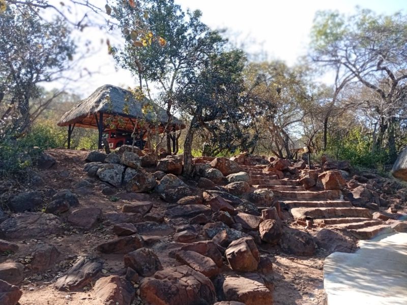 A natural garden setting featuring a thatched gazebo, stone staircase, and rugged rock landscaping within a wooded environment.