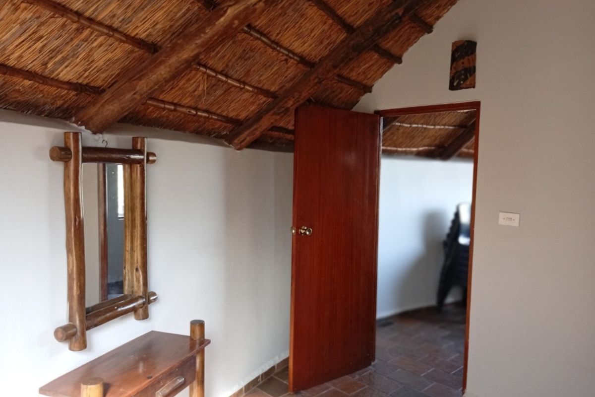 A bedroom featuring a rustic thatched roof with exposed timber rafters and tiled flooring, leading into an adjacent room through a wooden door.