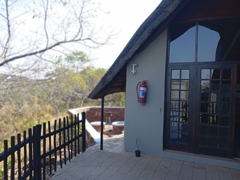 A covered patio area featuring a thatched roof, paved flooring, and timber-framed glass doors, overlooking a natural bushveld landscape.