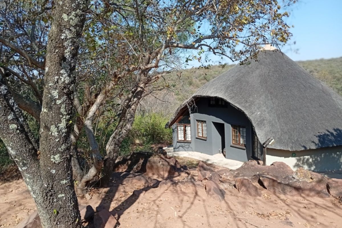 A charming cottage with a traditional thatch roof, set in a natural rocky landscape with mature trees.