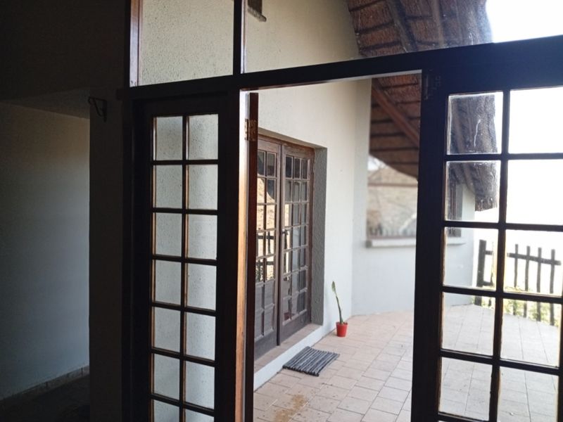 A covered patio area featuring tiled flooring, timber-framed glass doors, and a traditional thatched roof structure.