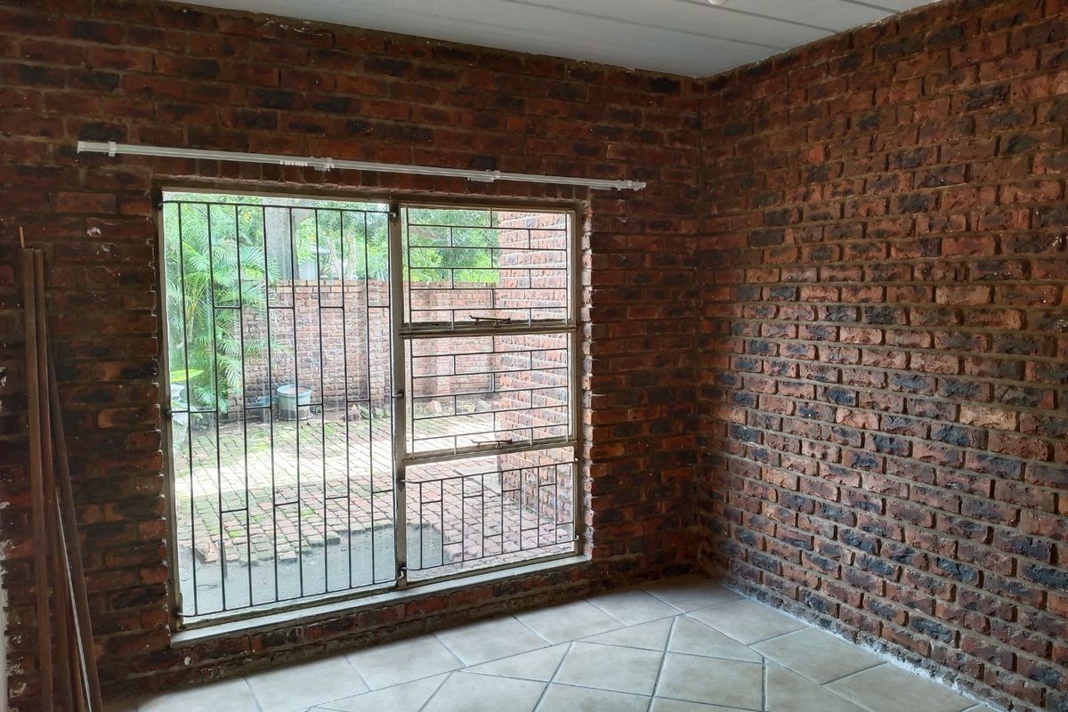An empty living area featuring exposed brick walls, tiled flooring, and a large window with security bars overlooking a rear courtyard.