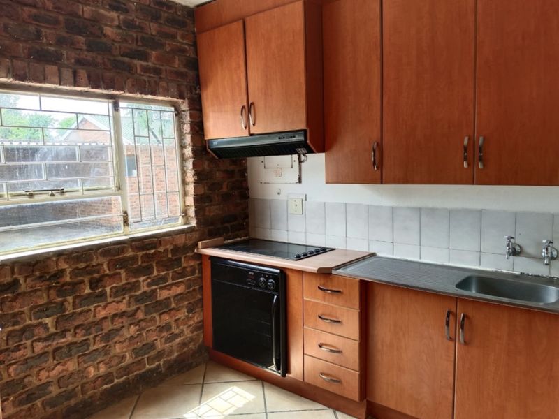 A dated kitchen featuring exposed brick walls, laminate cabinetry, built-in oven, electric hob, and a stainless steel sink with tiled splashback.