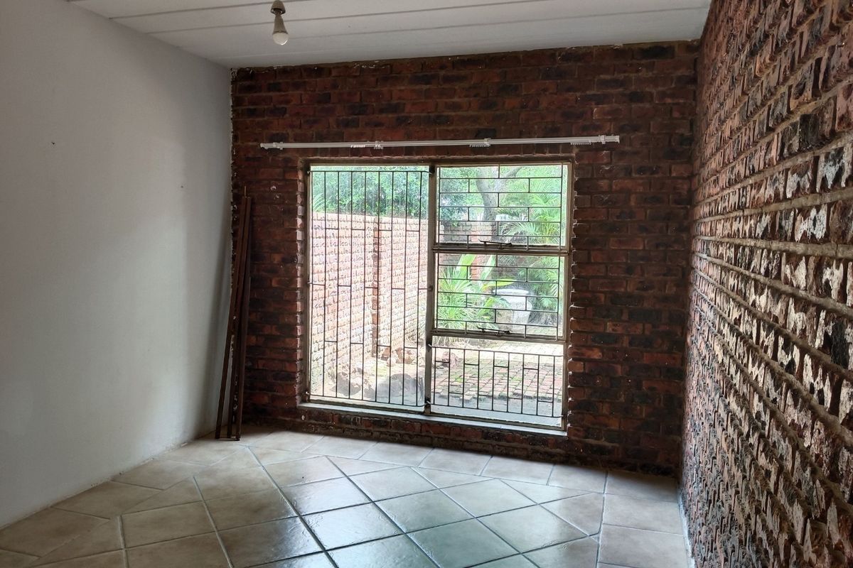 An empty bedroom featuring exposed brick walls, tiled flooring, and a window with security bars overlooking the exterior area.