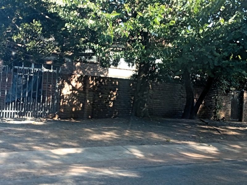 A street-level view of the property boundary featuring a brick wall, mature trees, and a metal security gate.