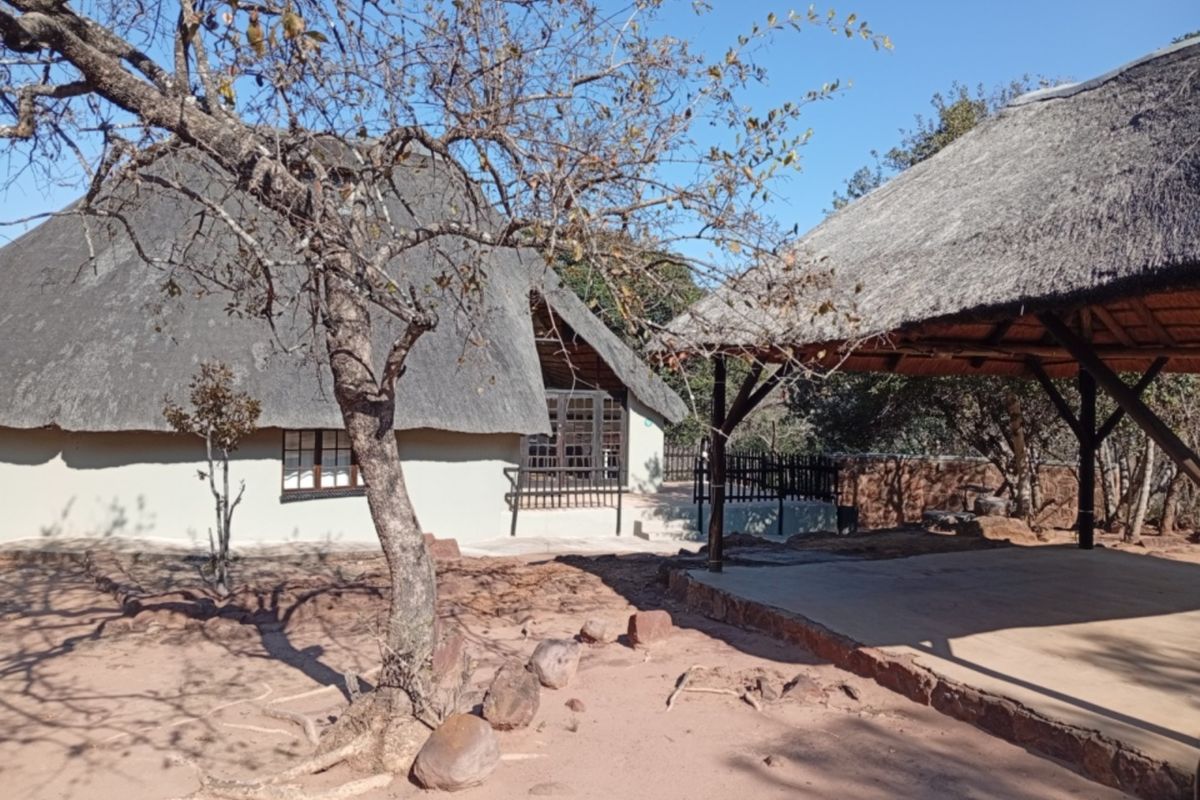 Exterior view of a rustic dwelling featuring a traditional thatch roof and an adjacent covered outdoor patio area set in a natural landscape.