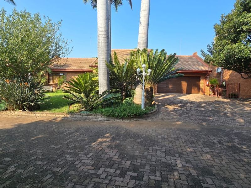 Front view of a brick townhouse featuring a paved driveway, attached garage, and established garden with mature palm trees.