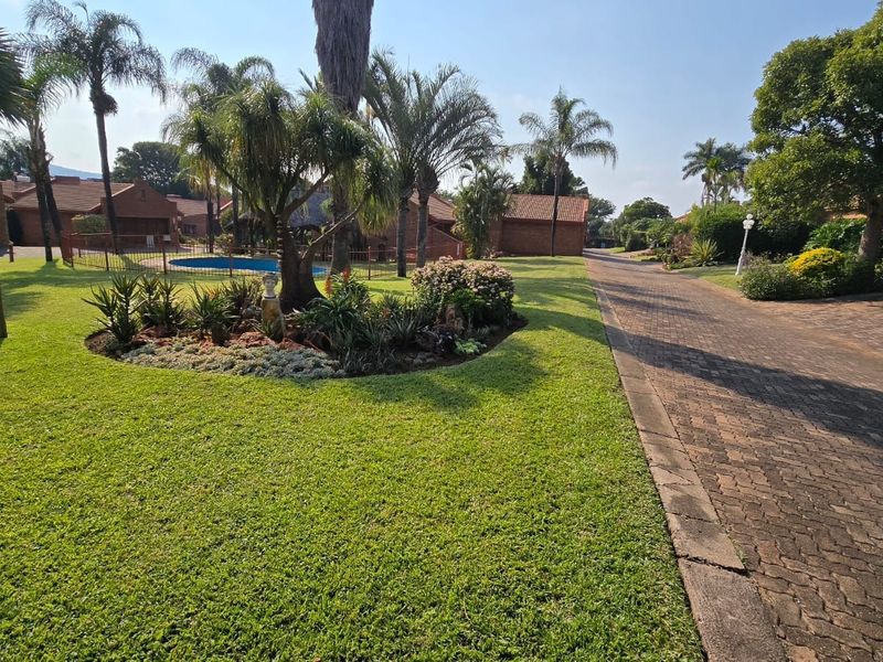 A well-maintained communal garden area featuring a paved driveway, lush landscaping, and a swimming pool within the townhouse complex.