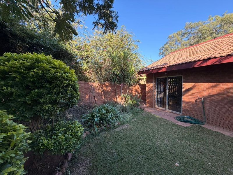 A private rear garden featuring a brick townhouse exterior, a small lawn, established landscaping, and a sliding door with security gate access.