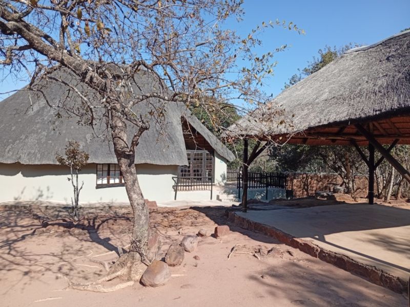 Exterior view of a rustic dwelling featuring a traditional thatch roof and an adjacent covered outdoor patio area set in a natural landscape.