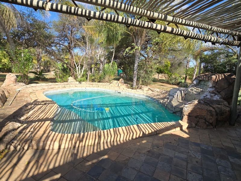 Outdoor pool area featuring a rock-accented swimming pool, paved patio, and a rustic timber pergola, set within a natural garden landscape.