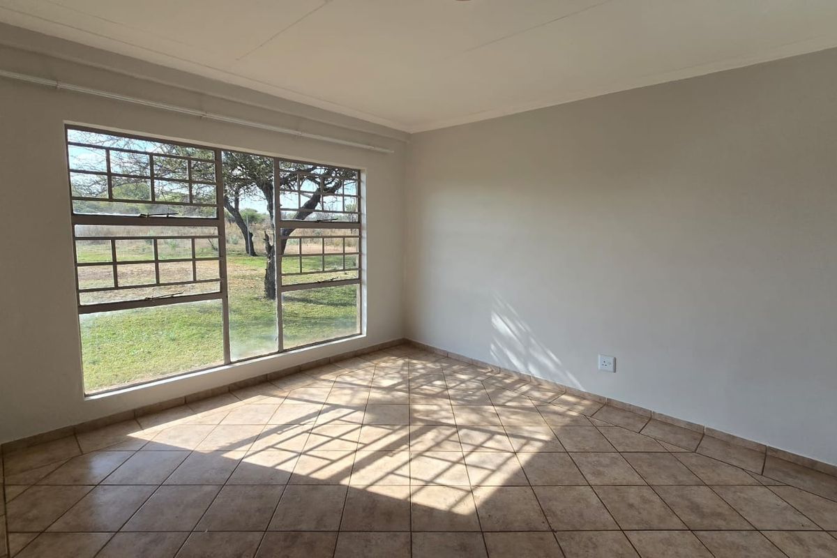 A bright, empty bedroom featuring neutral walls, tiled flooring, and a large window with security bars overlooking the garden.