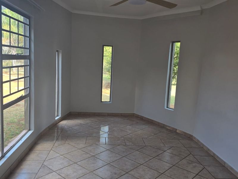 A bright, empty living area featuring neutral tiled flooring, a ceiling fan, and multiple vertical windows providing natural light and garden views.