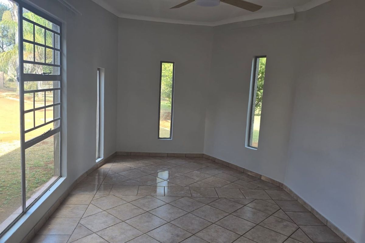 A bright, empty living area featuring neutral tiled flooring, a ceiling fan, and multiple vertical windows providing natural light and garden views.