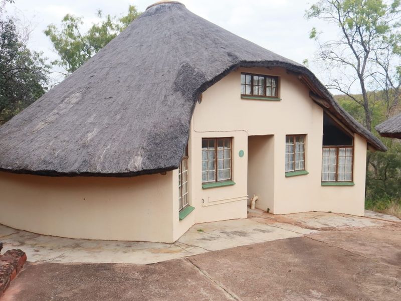A rustic cottage featuring a traditional thatched roof and plastered masonry walls, set within a natural landscape with a concrete entrance area.