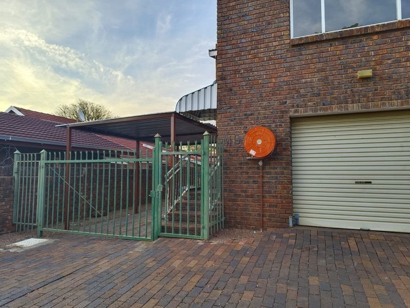 Front exterior view of the apartment featuring a brick facade, roller garage door, paved driveway, and a secure gated entrance with a covered patio area.
