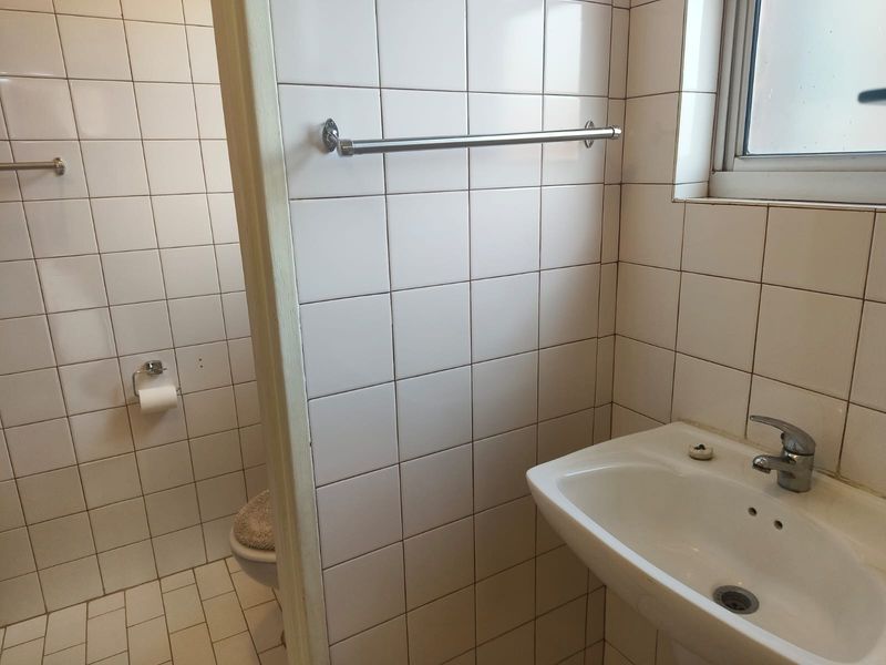 A functional bathroom featuring white wall tiles, a pedestal washbasin with a chrome mixer tap, and a window for natural light.