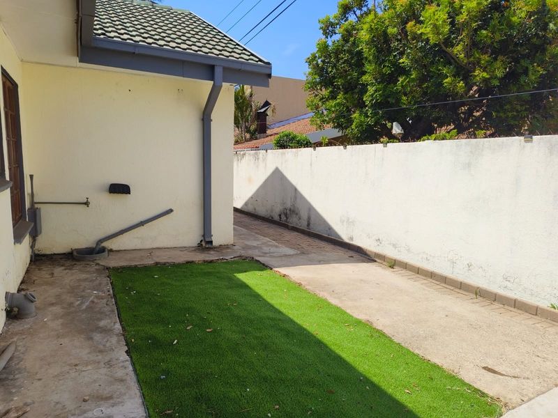 A side exterior view of the townhouse featuring a paved walkway, artificial lawn, and a boundary wall with some visible weathering.
