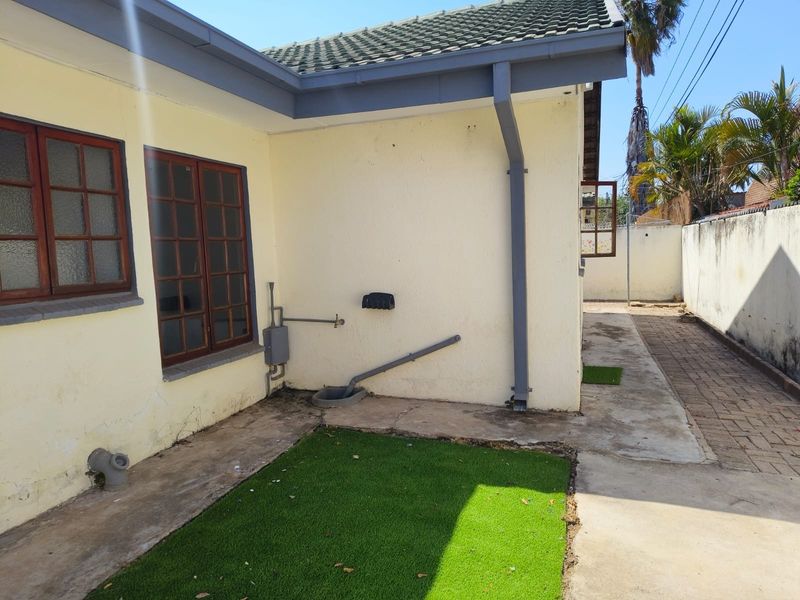 Rear exterior view of the townhouse featuring a small artificial lawn area, paved walkway, and boundary wall with visible wear to the exterior finishes.