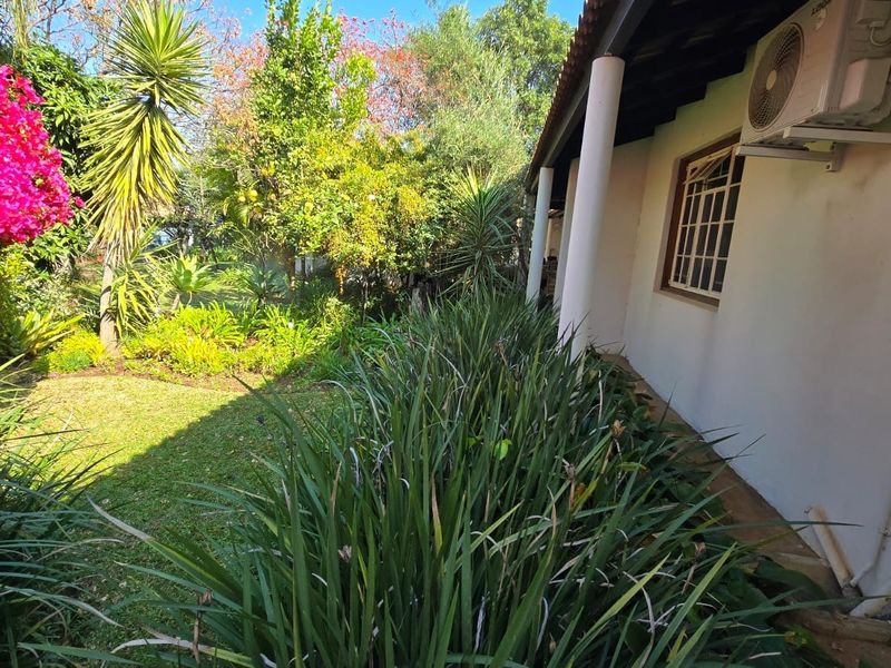A well-maintained garden area featuring lush landscaping, mature trees, and a covered patio space with an external air conditioning unit.