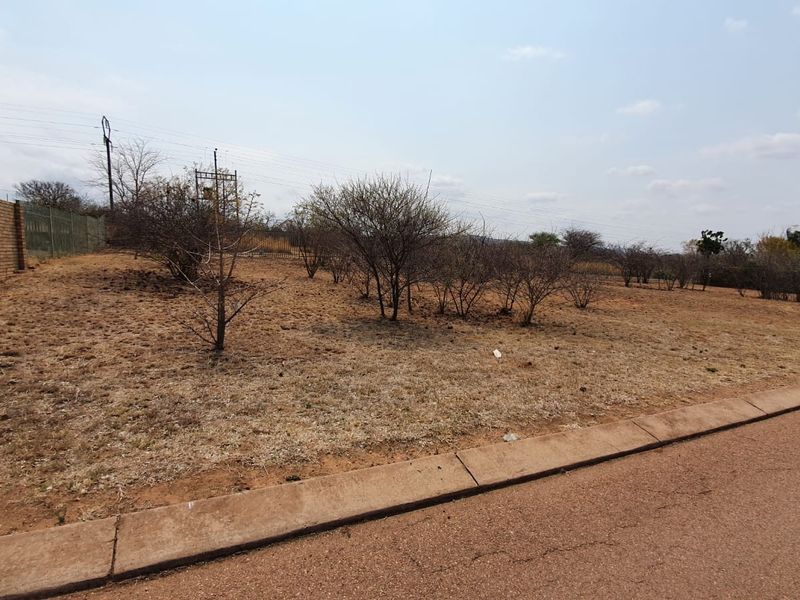 A vacant residential plot featuring dry grass, native shrubs, and established perimeter fencing, accessible from a paved road with existing utility infrastructure.